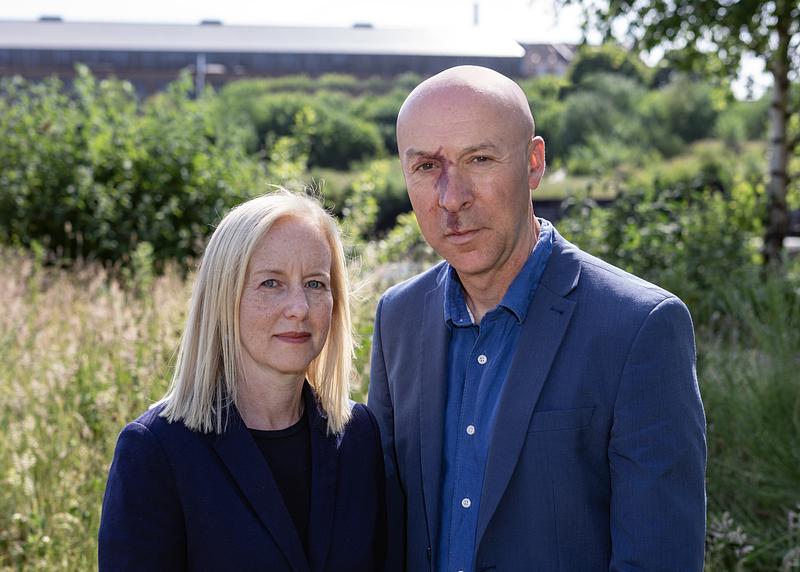A bald white man in a suit stands next to a white woman with blonde hair in a bob